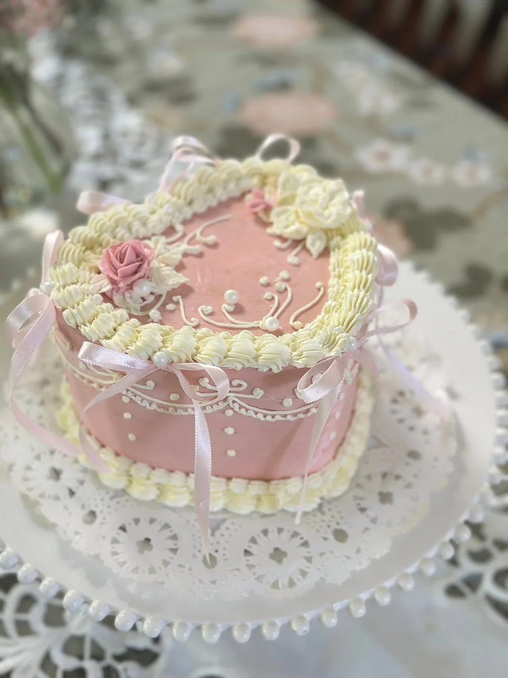 Our pink vintage heart cake, piped in cream with a sugar rose and pink ribbon bows, on a white lace doily