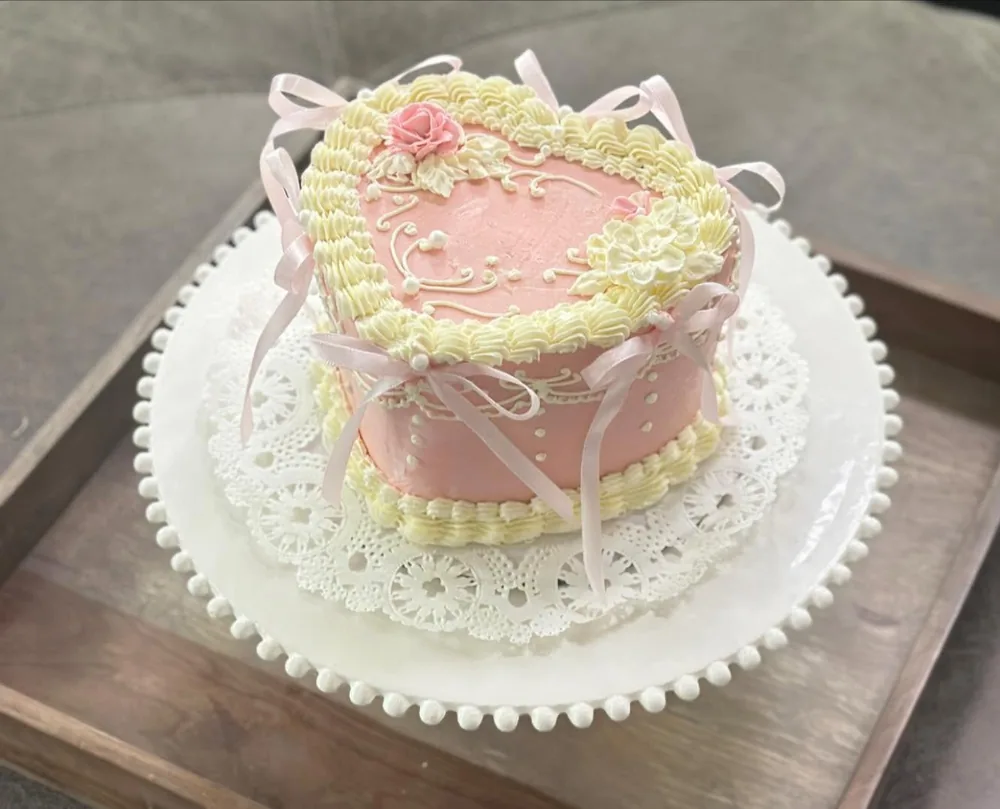 The pink vintage heart cake from above, with cream piping and ribbon bows, on a wooden tray