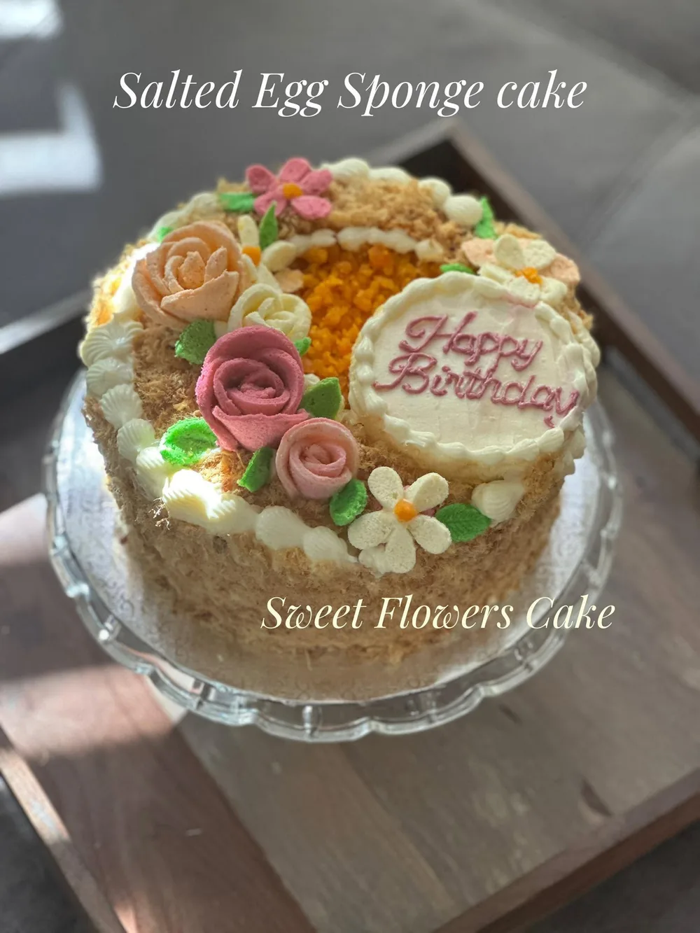 A salted egg sponge cake with buttercream roses and a Happy Birthday plaque, on a glass cake stand
