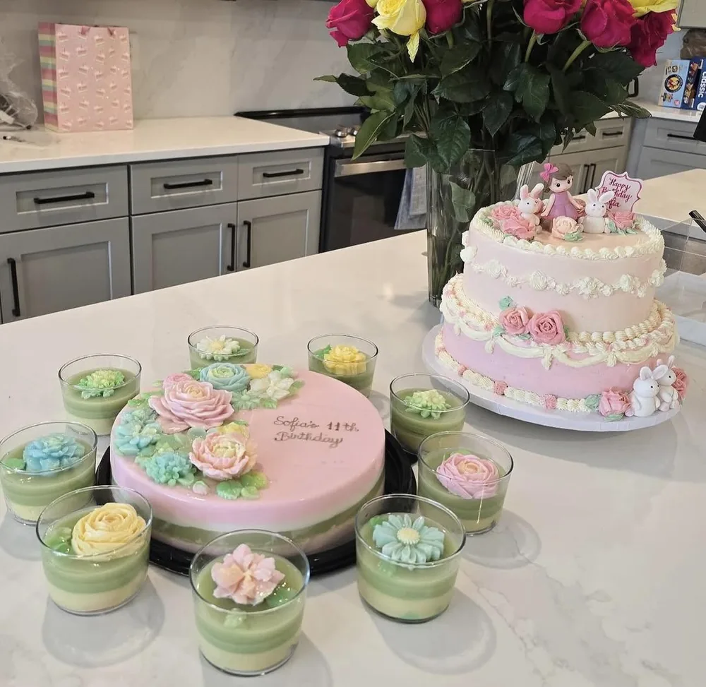 The Sofia jelly cake and jelly flower cups on a kitchen bench, beside a pink tiered cake and a vase of roses