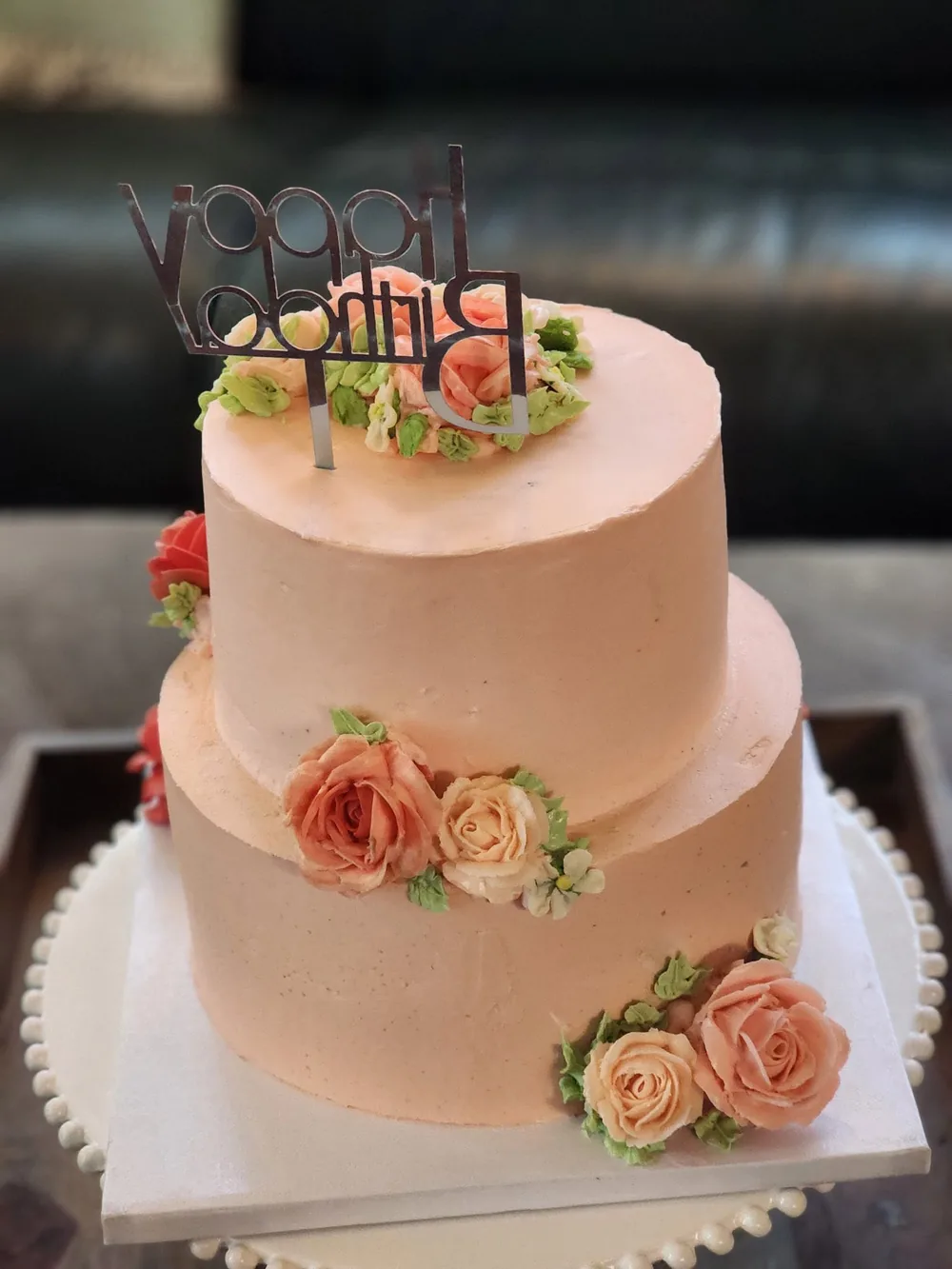 A two-tier peach cake with buttercream roses in coral and blush and a silver Happy Birthday topper